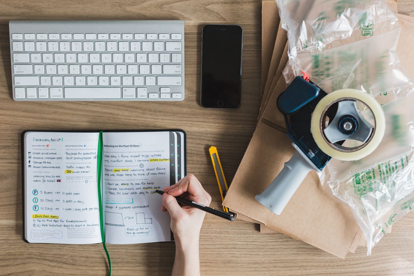 A notebook, laptop and packing tape on a work desk, seen from above
